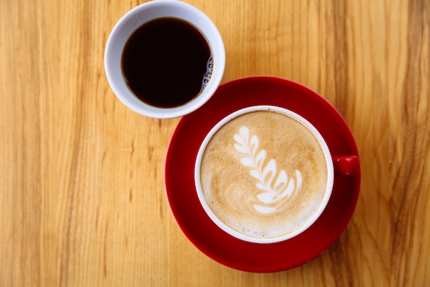 Кофе в красной чашке с молочной пеной и латте-арт и свежезаваренный кофе в белой чашке на столе.