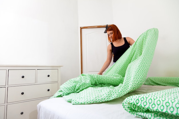 Â крупным планом молодая женщина в джинсах и черной футболке занимается домашними делами, убирает кровать с бело-зеленым геометрическим постельным бельем в спальне с белыми стенами и комодом.
