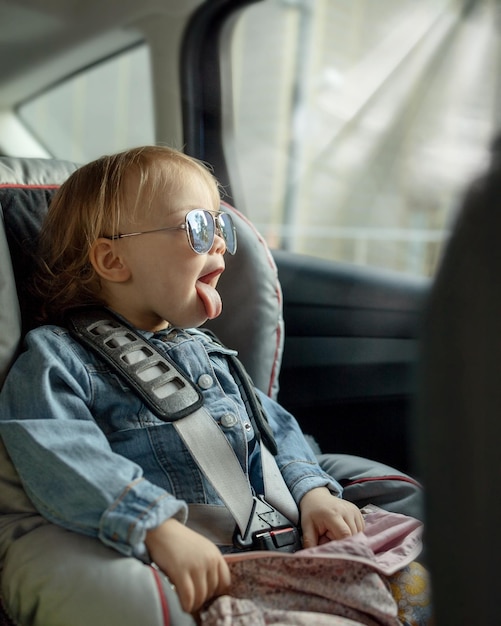 사진 엄마의 선글라스를 입은 아기가 차 좌석에 앉아서 창밖으로 혀를 고 있다.