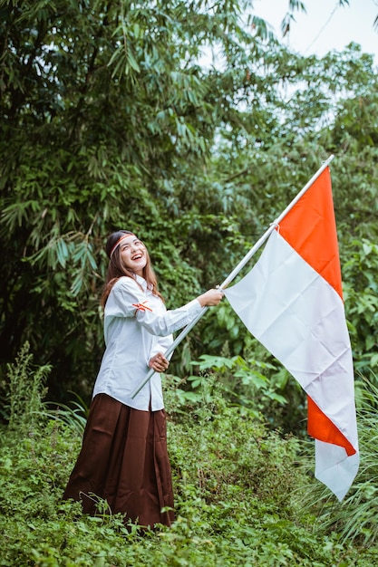 Фото Красивая азиатка в белой рубашке и коричневой юбке стоит и машет индонезийским флагом на руке.