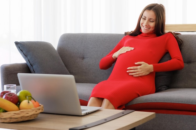 写真 美しい大きな妊婦がソファに座り、ノートパソコンを使ってビデオ通話で楽しく快適にチャットする 家族関係と感情ケアのコンセプト