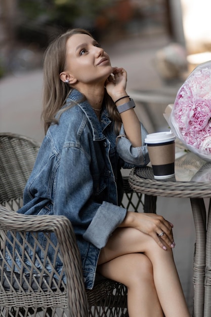 写真 カジュアルな服装をした美しい白人の女の子が通りのカフェに座り、美しい牡丹の花の隣でコーヒーを飲みます