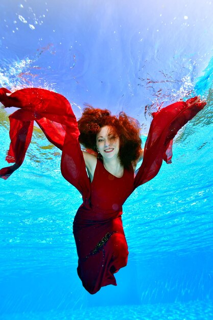 写真 赤い髪の美しい女性モデルが泳いでいて、赤いドレスを着て水中でポーズをとっています