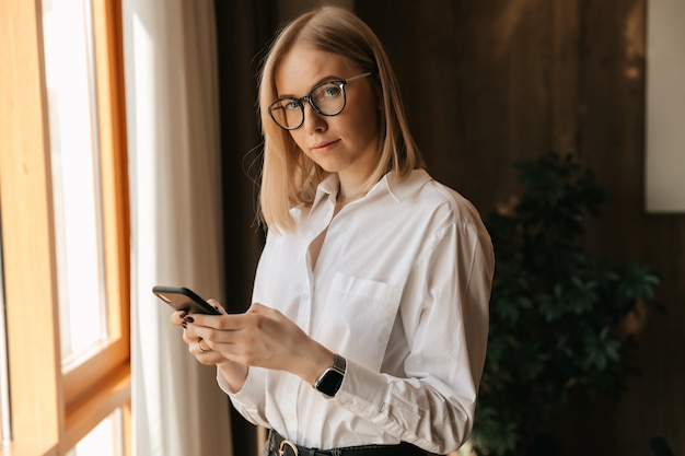 Фото Красивая девушка в очках стоит у окна в офисе в руках с телефоном, набирающим текст.