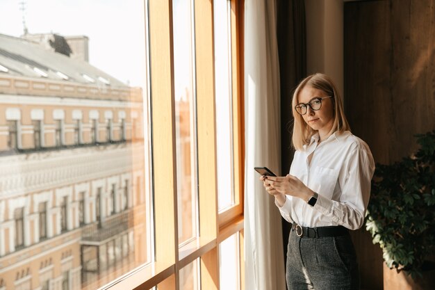Красивая девушка в очках стоит у окна в офисе в руках с телефоном, набирающим текст.