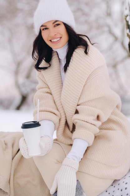 写真 湖の近くの雪に覆われた冬の森でお茶を楽しんでいるベージュのカーディガンと白い帽子をかぶった美しい少女