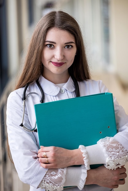彼女の手に本を持っている医療ガウンの美しい学生が病院の廊下に立っています医学生の概念女の子の肖像画