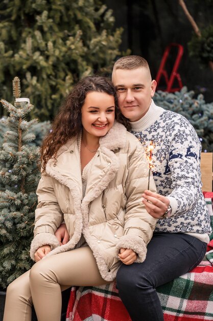 사진 화환 근처를 걷는 흰 옷을 입고 크리스마스 나무 근처에 앉아있는 아름다운 젊은 부부 행복한 남자와 여자 로맨스 크리스마스 축하 재미 사랑 흰 스웨터 새해 거리 밖