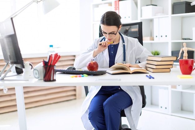 Красивая молодая девушка в белом халате сидит за компьютерным столом, держит ручку и работает с блокнотом и документами.