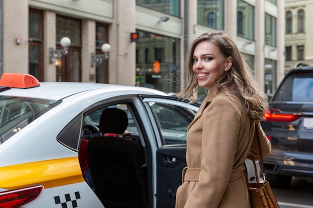 Фото Красивая молодая женщина садится в желтое такси в городе. улыбающаяся блондинка в бежевом пальто. путешествие и движение по мегаполису.