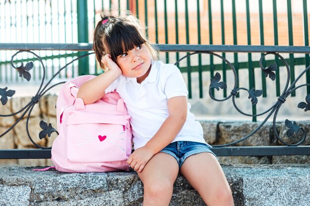 写真 ピンクのバックパックを身に着けている黒髪の少女は、学校の隣の石のベンチに座っています