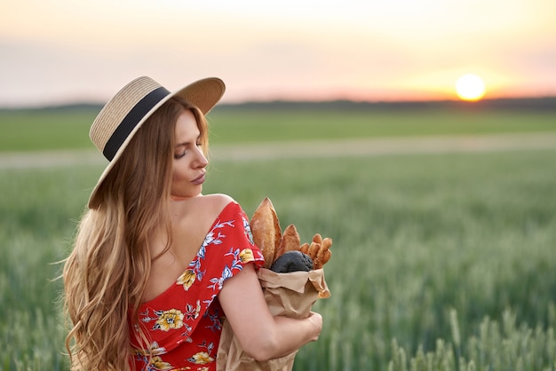 写真 赤いドレスを着た金髪と夕暮れ時の麦畑で彼女の手にフランスのパンと麦わら帽子