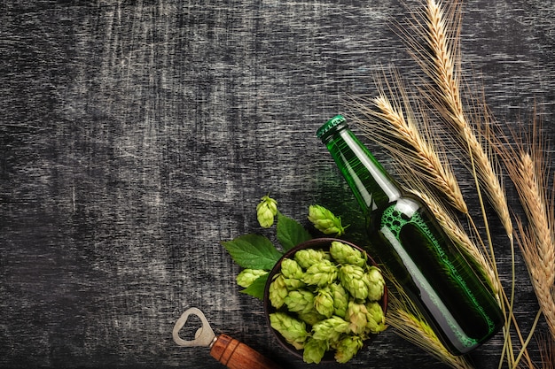 Фото Бутылка пива с зеленым хмелем, колоски и открывашка на черной поцарапанной доске
