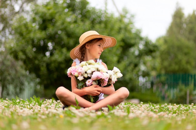 Букет розовых и белых пионов в руках девушки, сидящей на цветущем лугу.