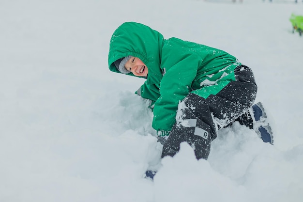 写真 緑のジャンプスーツを着た少年が雪に落ちた 少年が雪の中で遊ぶ 冬に外で遊ぶ子供