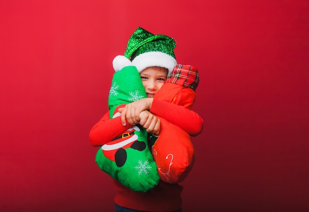 写真 ニットの赤いセーターとサンタ クロースの帽子をかぶった少年がプレゼント用のクリスマス ブーツを保持しています かわいい赤ちゃんはクリスマスと赤の背景に新しい種類の断熱材を待っています