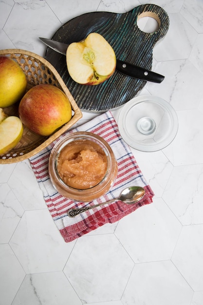 Фото Банка яблочного пюре и яблоки в плетеной тарелке на светлом фоне яблоко с ножом на деревянной разделочной доскевертикальная ориентация