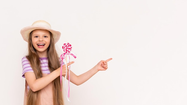 아름다운 소녀의 손에 있는 카라멜 롤리팝 여름 Sundress와 여름 모자를 쓴 아기는 검지 손가락으로 흰색 격리된 배경을 가리킵니다.