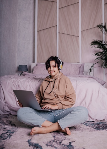 Кавказская девочка-подросток сидит возле кровати с ноутбуком и слушает музыку в наушниках