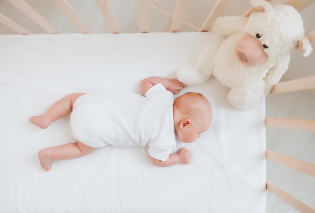 흰색 바디수트를 입은 매력적인 웃는 2개월 된 아기가 테디베어 옆에 있는 유아용 침대에 누워 있습니다.