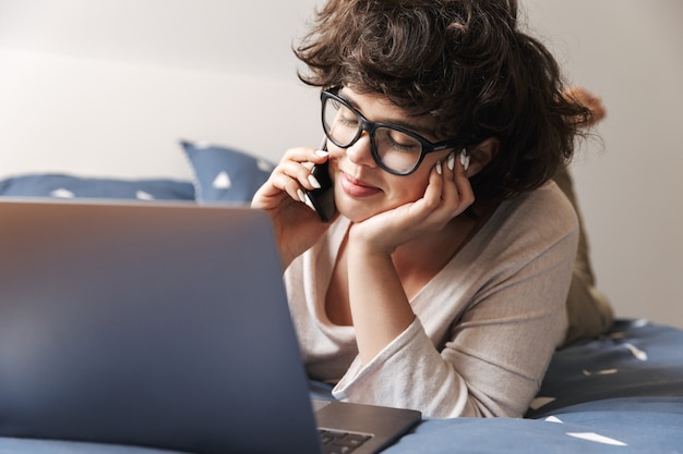 陽気なかわいい若い女性は、携帯電話で話しているラップトップコンピュータを使用してベッドの上に屋内で横たわっています。