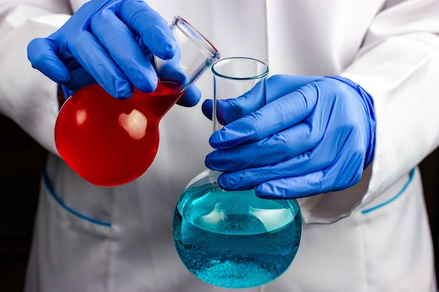 写真 白衣と青い手袋をはめた化学者が、液体の入った2つの化学フラスコを持っています