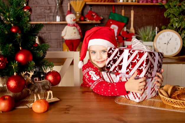 빨간 공이 있는 크리스마스 트리 옆 어두운 부엌에서 선물을 들고 산타 모자를 쓴 어린 소녀, 새해와 크리스마스의 개념