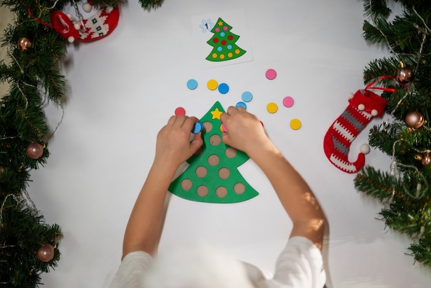 写真 頭に新年の帽子をかぶった白いtシャツを着た子供の女の子が、おもちゃのオリジナルのクリスマスツリーソーターをカットします。クリスマスの伝統の概念。クリスマスにモンテッソーリの細かい運動技能の発達。