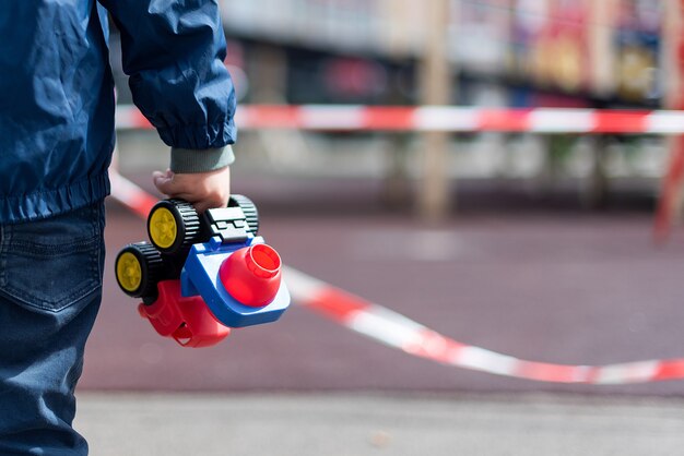 赤いリボンで囲われた遊び場の上に立っている子供がおもちゃの車を手に持っている