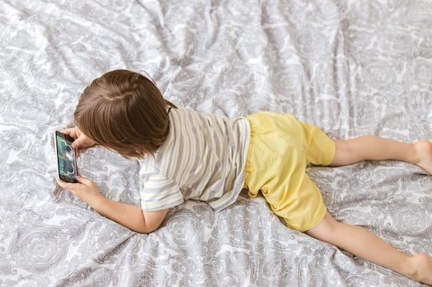Фото Милый мальчик лежит на кровати в спальне и играет с телефоном смартфона