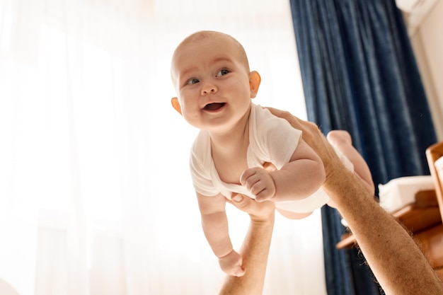 写真 父親の伸ばした腕の上で、明るい雰囲気の床に横たわるかわいいうれしそうな笑顔の赤ちゃん