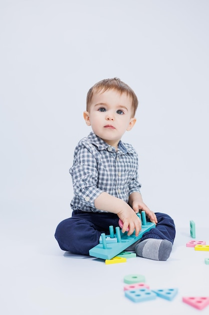 Фото Симпатичный маленький мальчик в клетчатой рубашке сидит на белом фоне и играет с деревянными развивающими игрушками экологические деревянные игрушки для детей