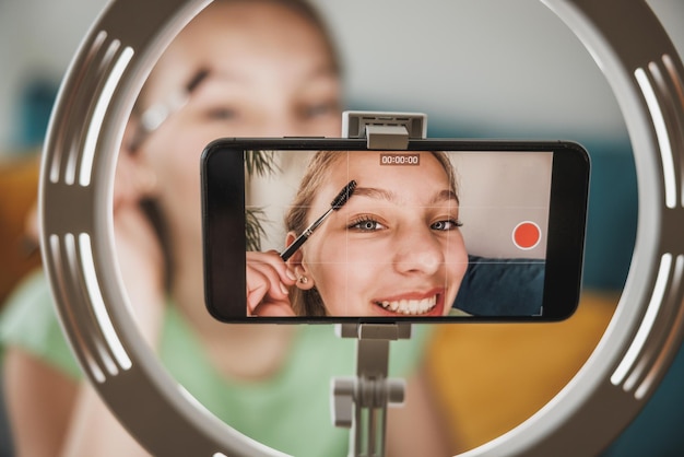 Симпатичная девочка-подросток-блогер в прямом эфире транслирует уроки макияжа для своих подписчиков через смартфон у себя дома.