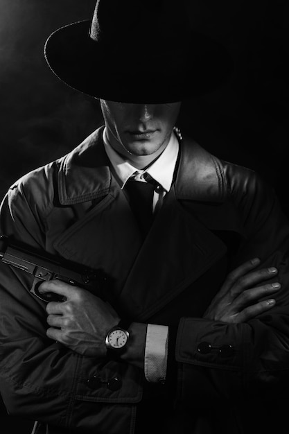 Фото Темный силуэт мужчины-детектива в пальто и шляпе с пистолетом в руках в стиле нуар голова опущена драматический нуарный портрет в стиле детективов 1950-х