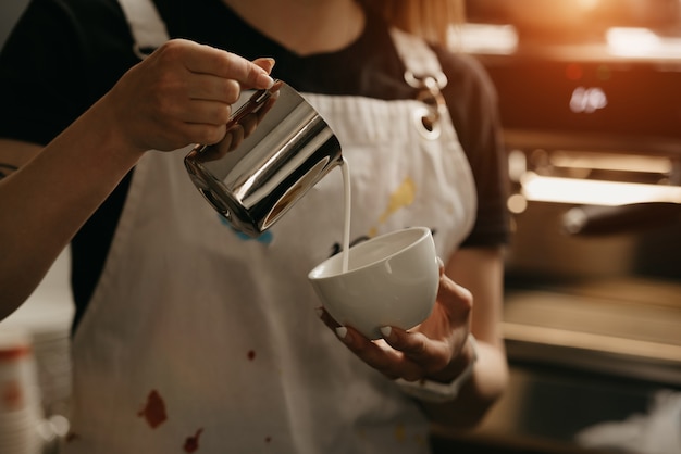 Фото Женщина-бариста с металлической кружкой молока изготавливает латте-арт на кофе в кафе. бариста наливает молоко узкой струйкой, чтобы вечером не разбить сливки эспрессо.