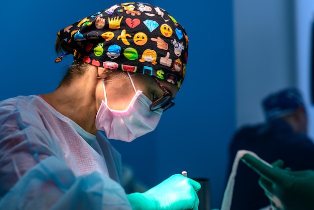 Фото Женщина-хирург стоит в операционной в забавной хирургической шапочке во время операции на синем фоне