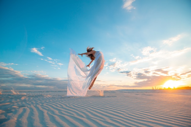 Девушка в летнем белом платье танцует и позирует в песчаной пустыне на закате