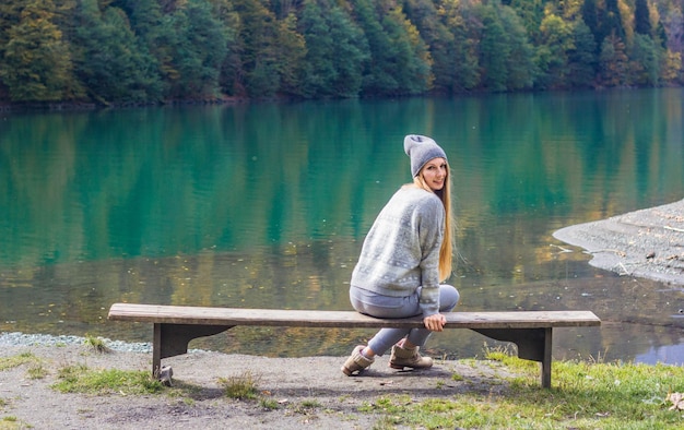 写真 帽子をかぶった女の子が秋の森と湖を背景にベンチに座っています