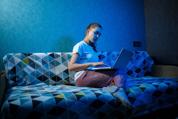 Девушка в белой футболке сидит дома на диване с ноутбуком на коленях и смотрит в экран. читает новую важную информацию. поиск новостей