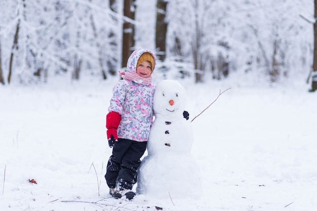 写真 冬の森の少女が雪だるまの隣に立っている