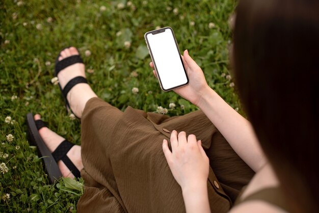 사진 올리브 드레스를 입은 소녀가 푸른 잔디에 앉아 그녀의 손에 흰색 화면이있는 전화를 보유하고 있습니다.
