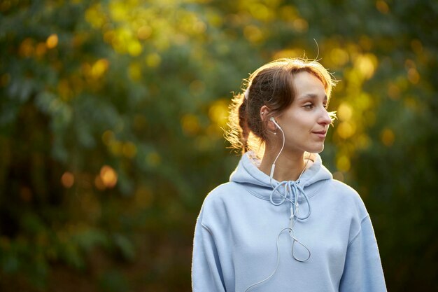 写真 青いパーカーを着たヨーロッパ風の女の子がヘッドフォンで市内の公園で音楽を聴く