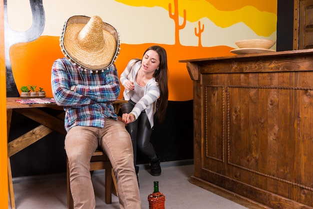 Девушка хочет украсть кошелек у мужчины, который напился и заснул, сидя на стуле и закрыв лицо сомбреро в мексиканском баре