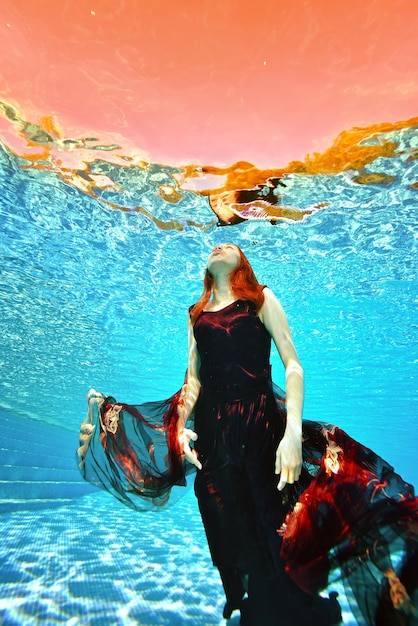 写真 赤い髪の少女が赤いドレスを着てプールの底から水面、そして太陽に浮かんでいます