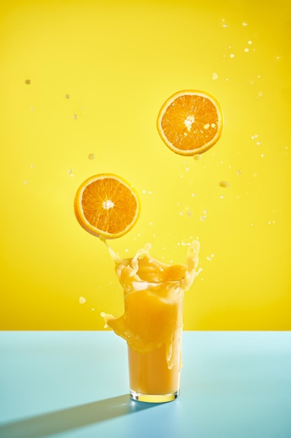 Фото Стакан брызг апельсинового сока и летающие апельсины на желтом фоне