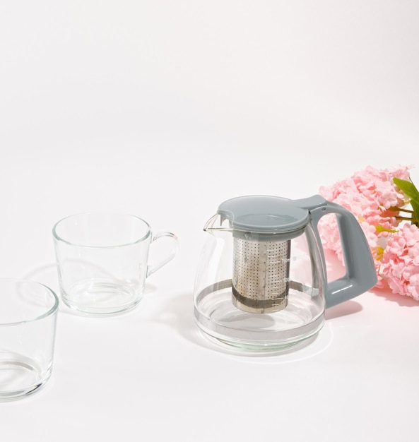 写真 グレーのキャップが付いたガラスのティーポットがテーブルの上に立っています 2 つの空のガラスのカップとピンクの花
