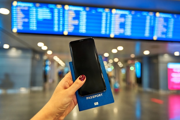 写真 空港の案内板を背景に、海外旅行用のスマートフォンとパスポートを手に持っています。