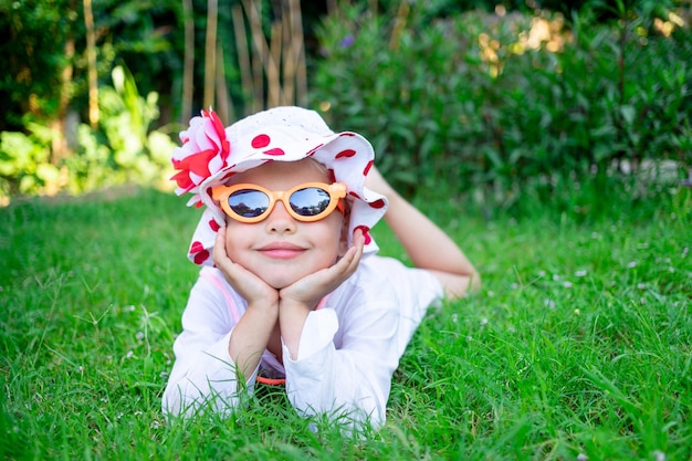 행복하고 즐거운 어린 소녀는 선글라스를 끼고 잔디밭의 푸른 잔디에 누워 있고 여름에는 파나마 모자를 쓰고 웃는다