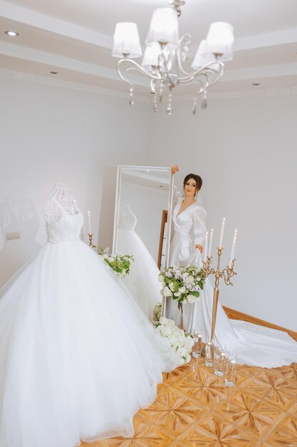 Фото Счастливая невеста готовится к своей роскошной свадьбе в гостиничном номере с свадебным платьем на манекене поблизости портрет женщины с модным макияжем волос и улыбкой в халате