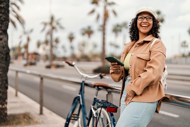 写真 幸せな世代の女の子が自転車の近くで携帯電話を手にして通りに傾いている